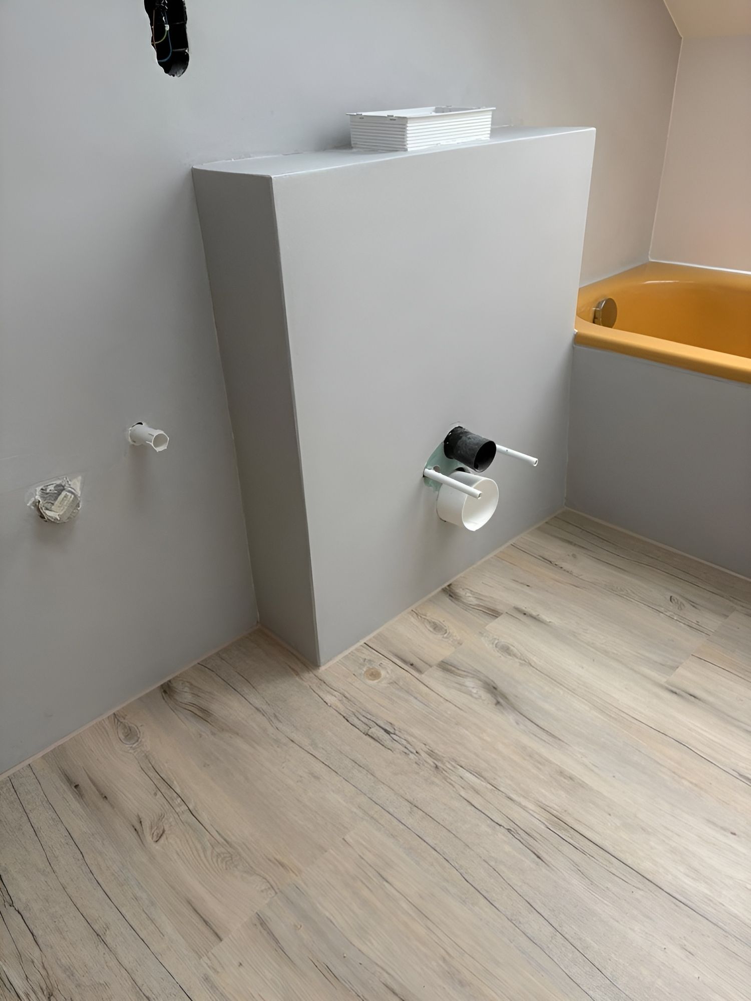 Eine teilweise fertiggestellte Badezimmerecke mit einer grauen Wand mit Sanitärarmaturen und einer gelben Badewanne. Heller Holzfußboden sichtbar.