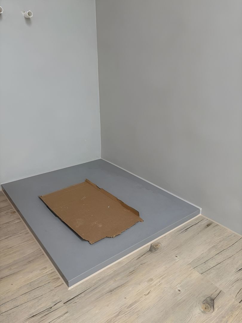 Ein minimalistisches Eckzimmer mit hellgrauen Wänden und einem grauen Podest, auf dem ein abgeflachter Pappkarton steht.