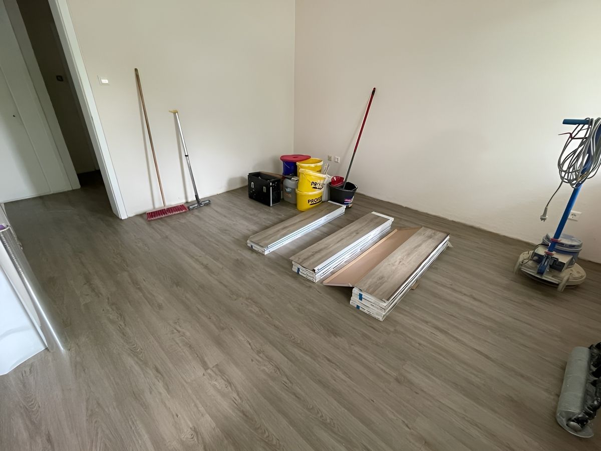 Ein teilweise eingerichteter Raum mit Holzfußboden, Reinigungsmitteln und Stapeln von Bodenbelägen, die für die Verlegung bereitstehen.