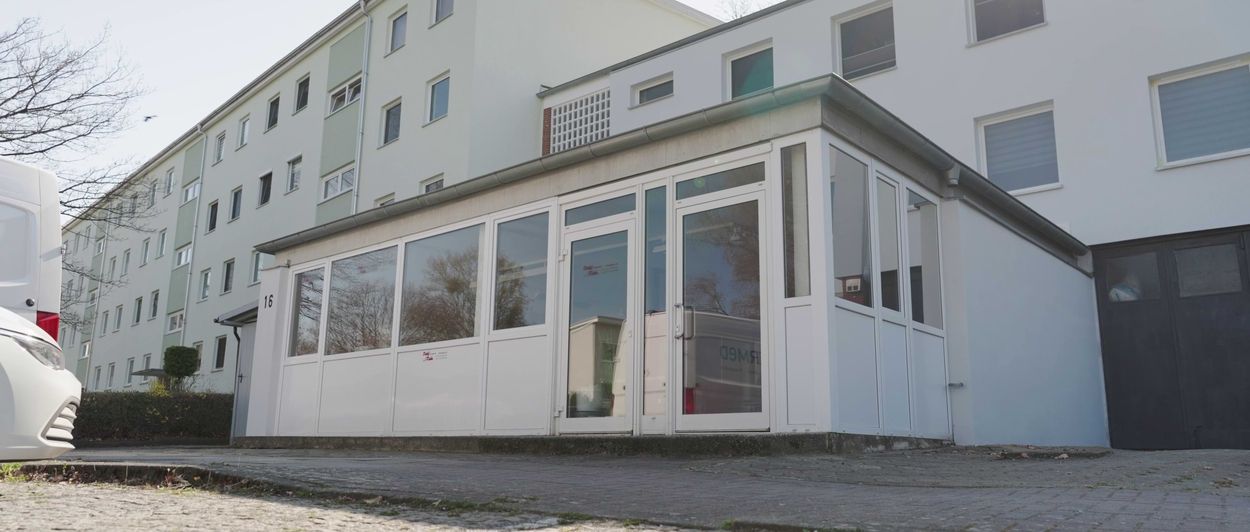 Ein weißer Wintergarten mit großen Glasfenstern ist an ein Wohnhaus angebaut, in dessen Nähe ein weißes Fahrzeug geparkt ist.