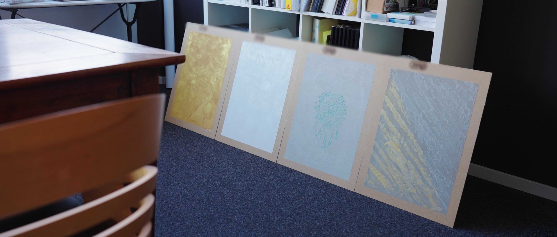 Vier Kunstwerke, die auf einem Holztisch vor einer dunklen Wand ausgestellt sind und verschiedene Texturen und Farben, darunter Gold und Hellblau, zeigen.